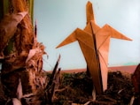 An origami figure crafted from brown paper is standing upright on dark soil, its pointed wings outstretched. It is positioned next to the textured, fibrous surface of a plant stub, highlighting the contrast between organic elements and the handcrafted paper object. The background is a muted blue, adding a calm atmosphere.