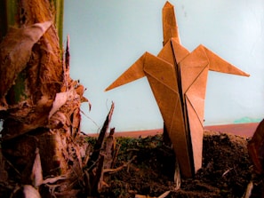 An origami figure crafted from brown paper is standing upright on dark soil, its pointed wings outstretched. It is positioned next to the textured, fibrous surface of a plant stub, highlighting the contrast between organic elements and the handcrafted paper object. The background is a muted blue, adding a calm atmosphere.