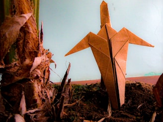 An origami figure crafted from brown paper is standing upright on dark soil, its pointed wings outstretched. It is positioned next to the textured, fibrous surface of a plant stub, highlighting the contrast between organic elements and the handcrafted paper object. The background is a muted blue, adding a calm atmosphere.