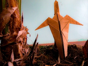 An origami figure crafted from brown paper is standing upright on dark soil, its pointed wings outstretched. It is positioned next to the textured, fibrous surface of a plant stub, highlighting the contrast between organic elements and the handcrafted paper object. The background is a muted blue, adding a calm atmosphere.
