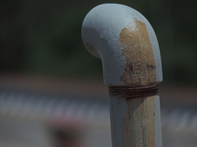 A rusted metal pipe with a curved end is displayed against a blurred background. The pipe shows signs of aging and corrosion, with noticeable rust patches and discoloration. The overall texture appears rough due to the corrosion.
