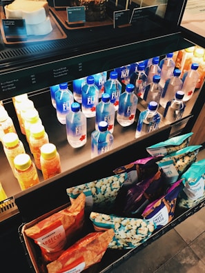 A refrigerated display case containing various beverages and snacks. The middle shelf features multiple bottles of Fiji Water alongside rows of yellow and orange juice bottles. Beneath this shelf, there is an assortment of packaged snacks, including bags of popcorn and potato chips. Some bakery items are visible on the top shelf.