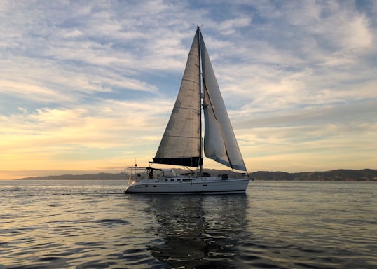 A calm sea at sunset with a small sailboat navigating gently under a sky filled with soft clouds.