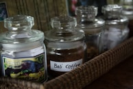 Several glass jars with lids are placed in a woven basket. One of the jars is labeled 'Bali Coffee' and contains brown coffee grounds.