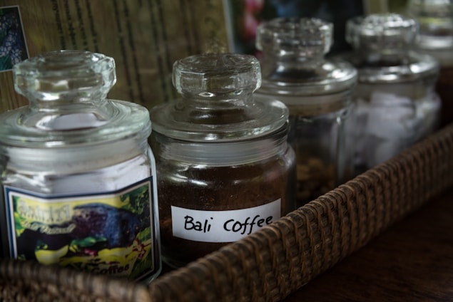 Several glass jars with lids are placed in a woven basket. One of the jars is labeled 'Bali Coffee' and contains brown coffee grounds.
