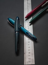 A group of three pens, each of different colors, lies on a dark surface with a transparent ruler beneath them. The pens are arranged in a loosely triangular pattern, and the ruler is placed at an angle, showing measurement units.