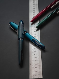 A group of three pens, each of different colors, lies on a dark surface with a transparent ruler beneath them. The pens are arranged in a loosely triangular pattern, and the ruler is placed at an angle, showing measurement units.