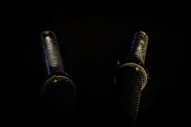 Two cylindrical, metallic objects with grooved patterns running lengthwise are positioned against a dark background. They appear to be bolts or screws with detailed threading visible, casting subtle reflections and shadows.
