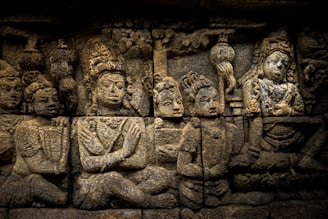 An intricately carved stone relief depicting a group of seated figures, likely of religious or cultural significance. The figures are adorned with headgear and jewelry, suggesting a historical or mythological context.