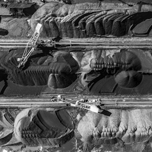Aerial view of a large industrial mining operation with massive excavators cutting through layers of earth and rock. The landscape is marked by deep ridges and patterns created by machinery, with heaps of excavated material forming a stark contrast against the machinery's metal structures.