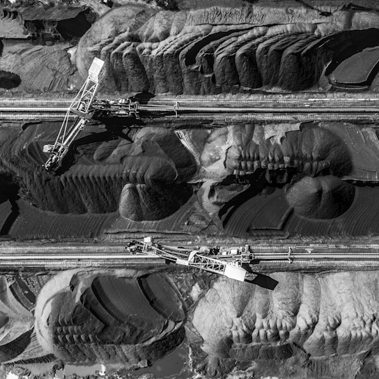 Aerial view of a large industrial mining operation with massive excavators cutting through layers of earth and rock. The landscape is marked by deep ridges and patterns created by machinery, with heaps of excavated material forming a stark contrast against the machinery's metal structures.