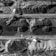 Aerial view of a large industrial mining operation with massive excavators cutting through layers of earth and rock. The landscape is marked by deep ridges and patterns created by machinery, with heaps of excavated material forming a stark contrast against the machinery's metal structures.