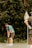 A golfer in a blue and white striped shirt and dark shorts is focusing on a short putt on a green. A flagstick is visible close by, while a lush, green background of trees adds a natural setting to the scene.