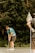 A golfer in a blue and white striped shirt and dark shorts is focusing on a short putt on a green. A flagstick is visible close by, while a lush, green background of trees adds a natural setting to the scene.
