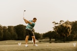 A person is playing golf on a lush green course, preparing to swing the golf club. The background features trees and an overcast sky. The golfer is wearing a striped polo shirt, shorts, and sports shoes.