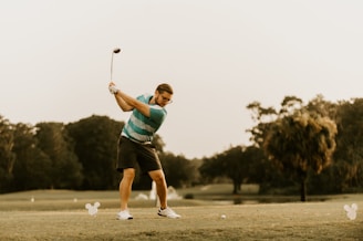 A person is playing golf on a lush green course, preparing to swing the golf club. The background features trees and an overcast sky. The golfer is wearing a striped polo shirt, shorts, and sports shoes.