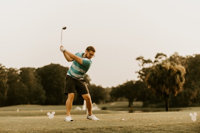 A person is playing golf on a lush green course, preparing to swing the golf club. The background features trees and an overcast sky. The golfer is wearing a striped polo shirt, shorts, and sports shoes.