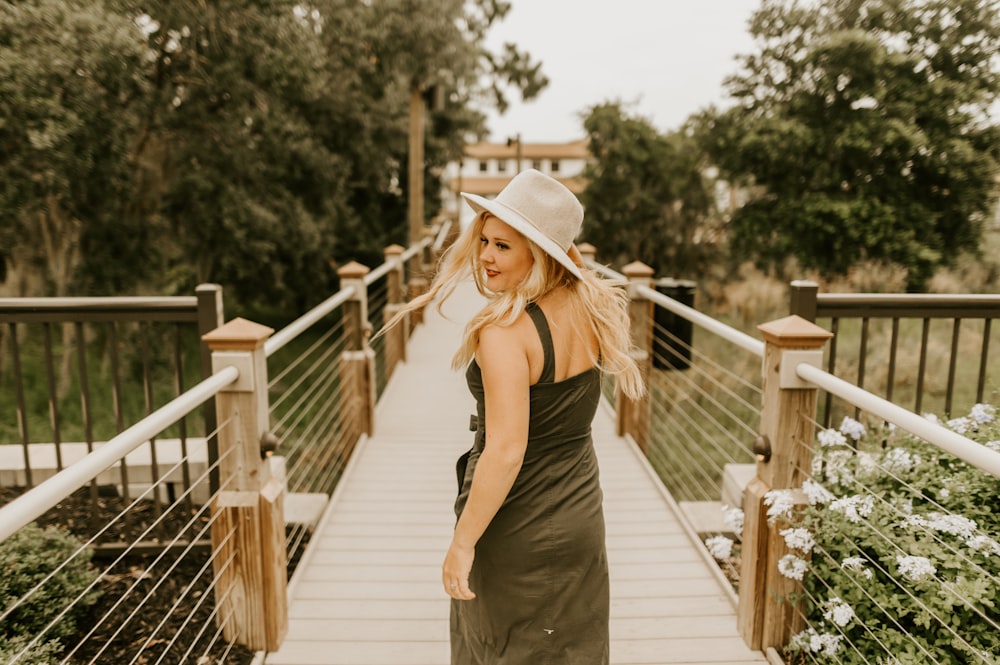 Frau in schwarzem Spaghettiträgerkleid und braunem Hut tagsüber auf Holzbrücke