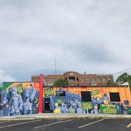 A vibrant mural on a building wall features various figures and signs with messages advocating for social justice. The artwork prominently includes phrases like 'Black Lives Matter,' and other calls for societal change. The imagery uses a mix of colors and geometric patterns as a backdrop, with figures wearing masks, highlighting themes of activism and awareness.