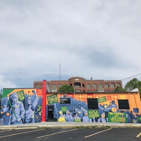 A vibrant mural on a building wall features various figures and signs with messages advocating for social justice. The artwork prominently includes phrases like 'Black Lives Matter,' and other calls for societal change. The imagery uses a mix of colors and geometric patterns as a backdrop, with figures wearing masks, highlighting themes of activism and awareness.