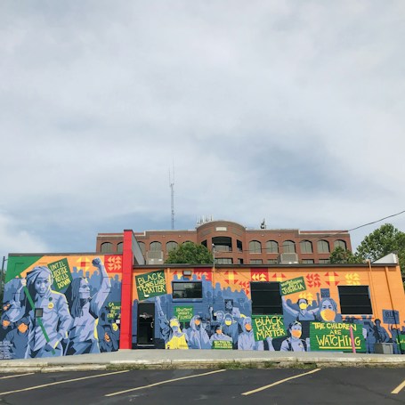 A vibrant mural on a building wall features various figures and signs with messages advocating for social justice. The artwork prominently includes phrases like 'Black Lives Matter,' and other calls for societal change. The imagery uses a mix of colors and geometric patterns as a backdrop, with figures wearing masks, highlighting themes of activism and awareness.