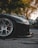 A close-up view of a black sports car's front left wheel and bumper, with a shiny, silver alloy rim set on a clean asphalt road. The car appears to be parked in an outdoor setting with a background of trees and other parked cars. The sunlight filters through the trees, creating a serene and somewhat dramatic ambiance.