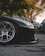 A close-up view of a black sports car's front left wheel and bumper, with a shiny, silver alloy rim set on a clean asphalt road. The car appears to be parked in an outdoor setting with a background of trees and other parked cars. The sunlight filters through the trees, creating a serene and somewhat dramatic ambiance.