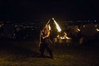 A performer is skillfully twirling a fire staff in a nighttime outdoor setting. The background includes the faint lights of a distant city and a small fire burning in metal drums. The scene is dimly lit, accentuating the fire and creating dramatic shadows.
