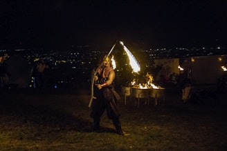 A performer is skillfully twirling a fire staff in a nighttime outdoor setting. The background includes the faint lights of a distant city and a small fire burning in metal drums. The scene is dimly lit, accentuating the fire and creating dramatic shadows.
