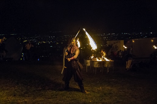 A performer is skillfully twirling a fire staff in a nighttime outdoor setting. The background includes the faint lights of a distant city and a small fire burning in metal drums. The scene is dimly lit, accentuating the fire and creating dramatic shadows.