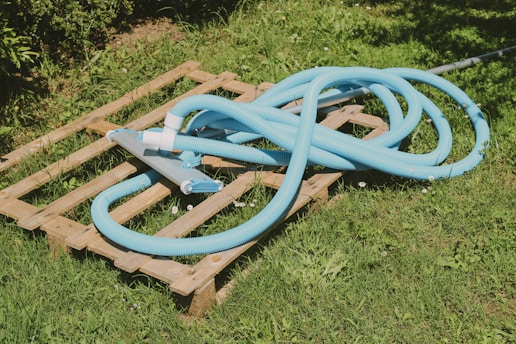 A coiled blue pool hose rests on a wooden pallet situated on grass. The sun casts shadows and highlights on the various textures of the grass, pallet, and hose. Small white flowers are scattered in the grass surrounding the pallet.