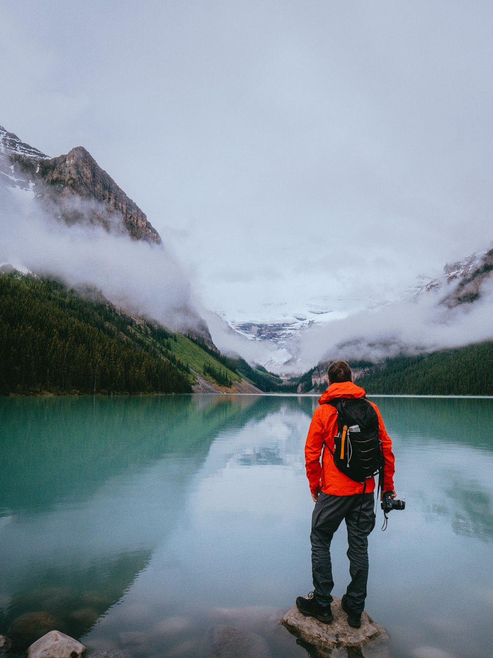 Hombre con chaqueta roja y negra y pantalones negros de pie en la roca cerca del lago durante el día