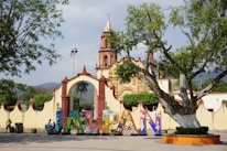 A vibrant town square features a large, colorful sign spelling out 'JALPAN' in front of an ornate, historical building. The structure is accented with red and cream colors with a bell tower reaching into the sky. The scene is framed by lush green trees, with a clear sky and distant hills providing a picturesque backdrop.