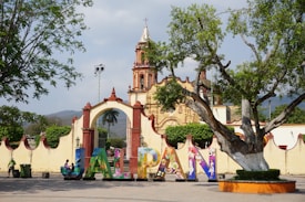 A vibrant town square features a large, colorful sign spelling out 'JALPAN' in front of an ornate, historical building. The structure is accented with red and cream colors with a bell tower reaching into the sky. The scene is framed by lush green trees, with a clear sky and distant hills providing a picturesque backdrop.