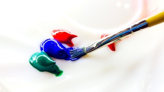 A close-up of a paintbrush resting on a surface with blobs of blue, red, and green paint. The bristles of the brush have some blue paint on them, indicating it's in the process of painting or blending colors.