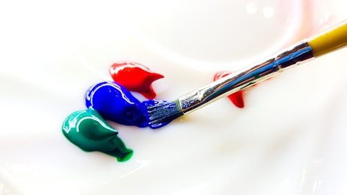 A close-up of a paintbrush resting on a surface with blobs of blue, red, and green paint. The bristles of the brush have some blue paint on them, indicating it's in the process of painting or blending colors.