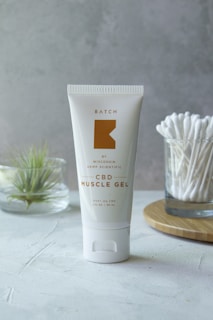 A tube of CBD Muscle Gel is placed upright on a light-colored surface. The tube is white with a minimalist design, featuring gold text and branding. In the background, there is a small glass container holding a plant and another glass filled with cotton swabs on a wooden tray.