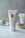 A tube of CBD Muscle Gel is placed upright on a light-colored surface. The tube is white with a minimalist design, featuring gold text and branding. In the background, there is a small glass container holding a plant and another glass filled with cotton swabs on a wooden tray.
