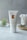 A tube of CBD Muscle Gel is placed upright on a light-colored surface. The tube is white with a minimalist design, featuring gold text and branding. In the background, there is a small glass container holding a plant and another glass filled with cotton swabs on a wooden tray.
