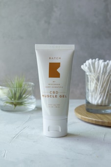 A tube of CBD Muscle Gel is placed upright on a light-colored surface. The tube is white with a minimalist design, featuring gold text and branding. In the background, there is a small glass container holding a plant and another glass filled with cotton swabs on a wooden tray.