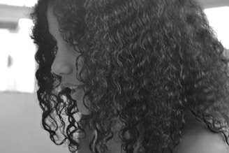 A black and white close-up profile view of a person with curly hair. The focus is on the texture and volume of the curls, with the background slightly blurred.