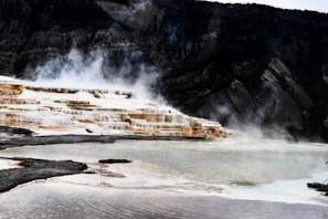 Steam rises from terraced hot springs against a backdrop of rugged mountains. The formations create a cascading effect, with various shades of brown, white, and golden hues. The foreground features rippling water reflecting the colors of the terraces.