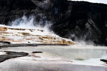 Steam rises from terraced hot springs against a backdrop of rugged mountains. The formations create a cascading effect, with various shades of brown, white, and golden hues. The foreground features rippling water reflecting the colors of the terraces.
