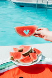 A hand holding a watermelon slice with a heart cutout above a pool