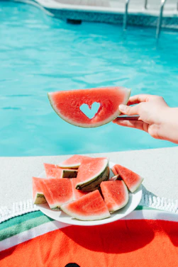 A hand holding a watermelon slice with a heart cutout above a pool