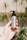 A hand with red nail polish holding a silver bottle of annmarie Sweet Sunrise Shampoo with a black pump dispenser. The background is slightly blurred, showing some greenery and a bright, sunny environment.