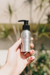 A hand with red nail polish holding a silver bottle of annmarie Sweet Sunrise Shampoo with a black pump dispenser. The background is slightly blurred, showing some greenery and a bright, sunny environment.