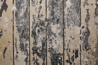Weathered wooden panels with peeling paint, displaying a rustic and distressed look. Various shades of beige and dark gray create a textured pattern across the surface.