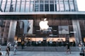 An exterior view of a large, modern retail store with a prominent glowing apple logo on the glass facade. Several people are walking in front of the store, which includes tall glass windows and signs indicating product displays inside.