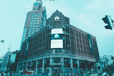 A large commercial building prominently displays the Huawei logo and name. The structure features modern architecture with vertical lines and a large digital screen advertising Huawei products. Several other high-rise buildings are visible in the background, and there is a busy street scene with people and traffic at the bottom of the image.